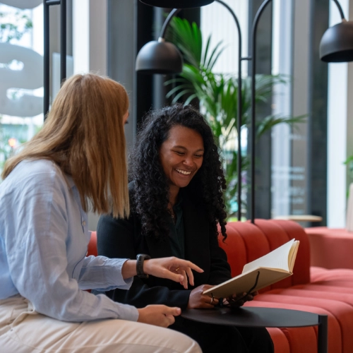 Two professionals discussing documents in a modern office lounge, representing careers and workplace collaboration